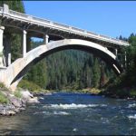 An art deco bridge spans a rural river with green trees on both sides.