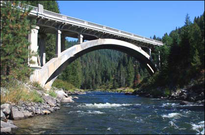 An art deco bridge spans a rural river with green trees on both sides.