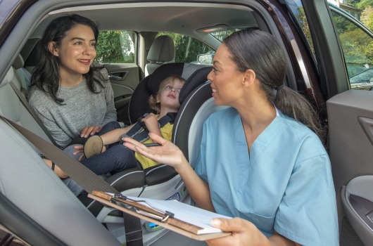 A nurse talks with a mother about child passenger safety while a young child sits in a car seat.