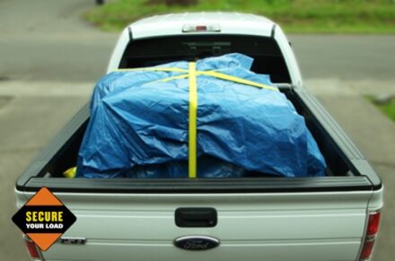 A white truck with cargo in the truck bed, the load is safely secured with a blue tarp and yellow straps.