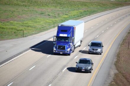 A blue semi truck and two cars driving on the interstate.