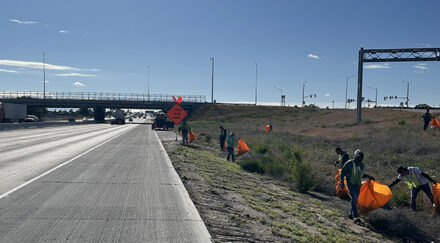 People with orange bags and safety vests pick up trash on the side of the highway.