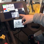 A hand points out the screens inside the cab of a snowplow, one of which shows the truck bed holding salt.