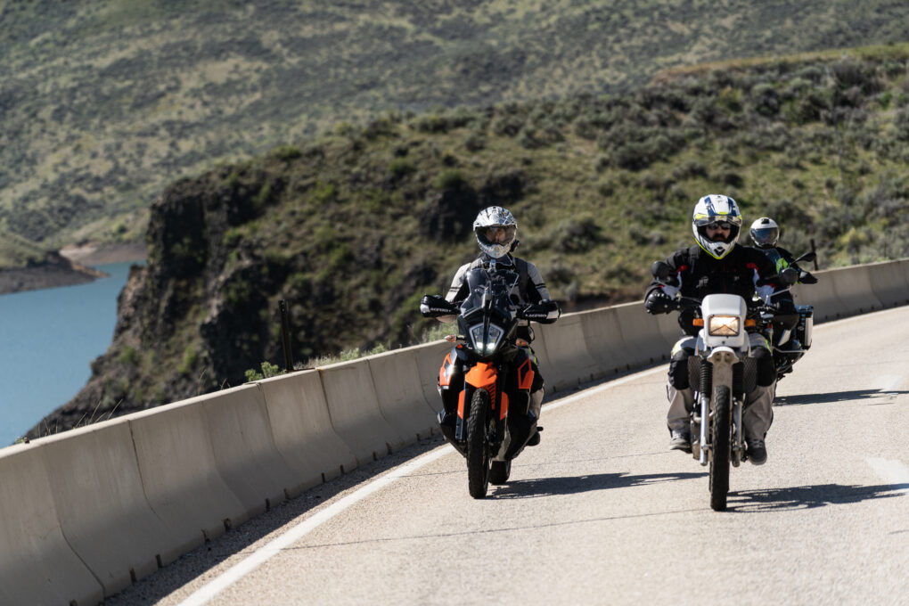 Two motorcyclists riding on a road. Hills and water are visible in the background.