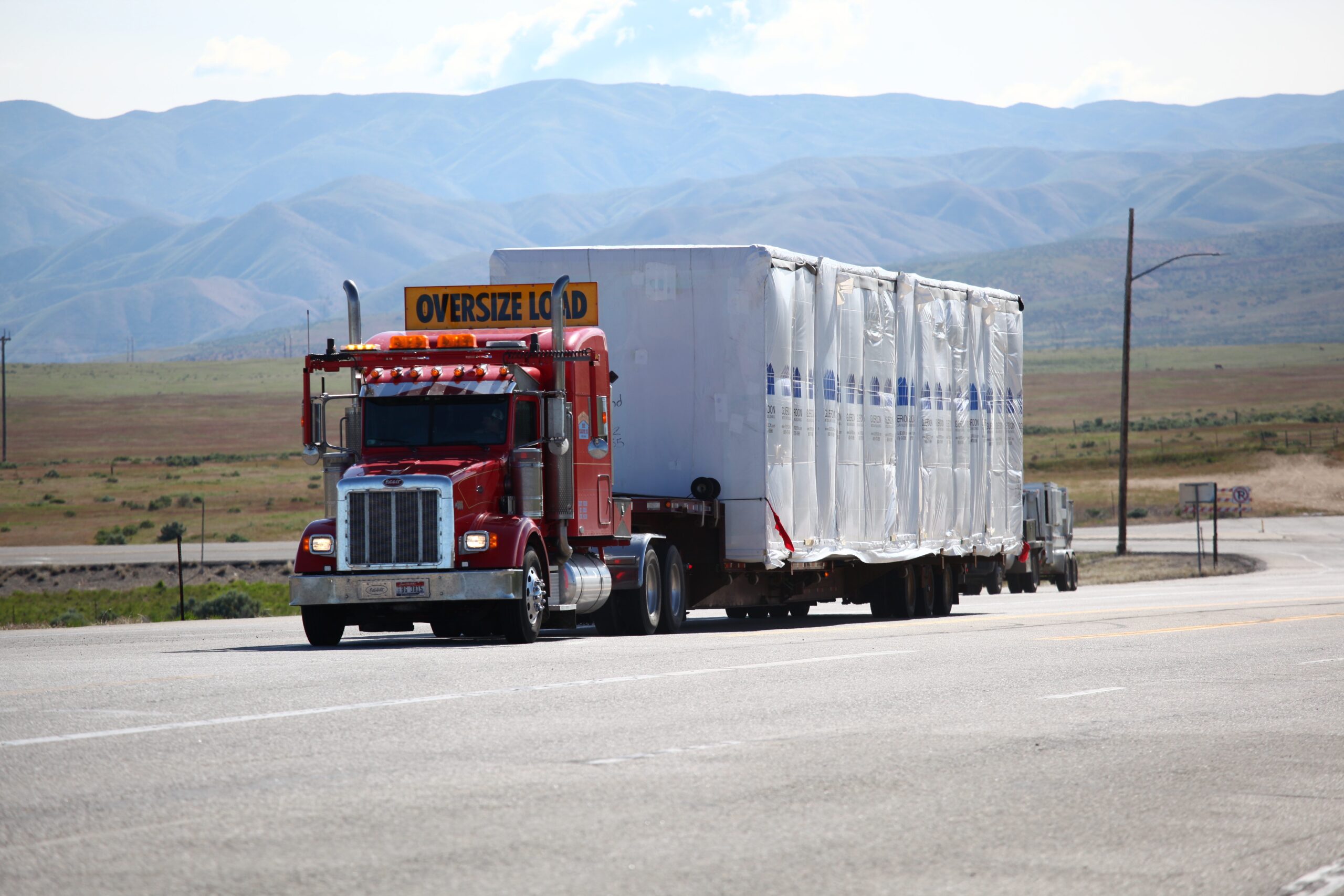 A red semi truck hauling an oversize load wrapped in white plastic.