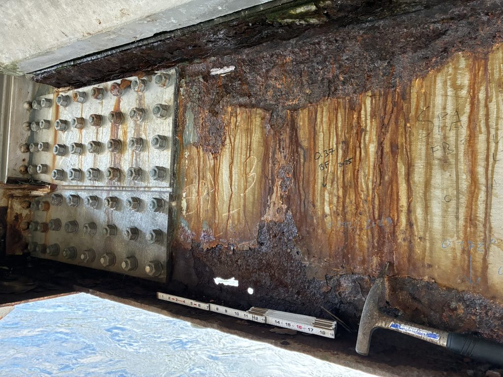 Close-up of significant rust and corrosion on a steel member of the East Kooskia Bridge, with rust streaking, exposed deteriorated metal, bolted reinforcement plates, measurement markings and a measuring stick visible.