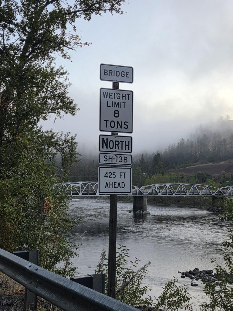 An 8-ton bridge weight limit warning sign stands along SH-13B with the East Kooskia Bridge visible across the Clearwater River in the background on a foggy morning.