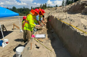 Three people working side by side to excavate dirt near a state highway.