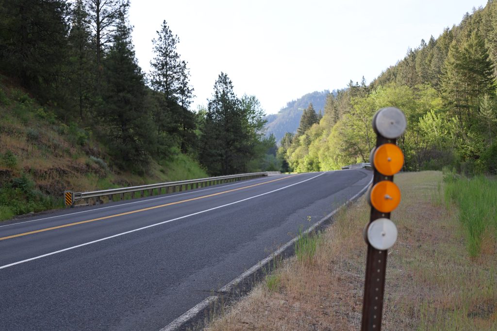 Rural highway curves through a forested canyon with roadside delineators in the foreground and mountains in the distance.
