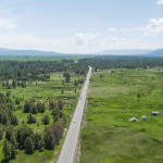 A two-lane highway stretches through a meadow with mountains in the background
