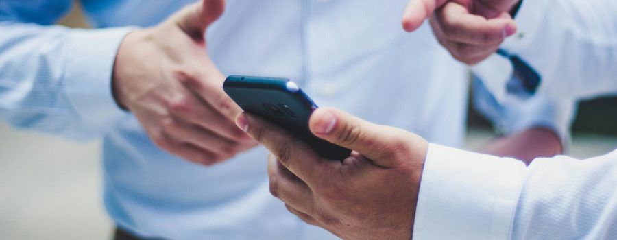 Two men standing together. One is holding a phone and pointing to the screen.