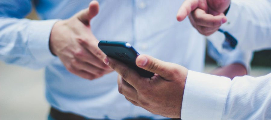 Two men standing together. One is holding a phone and pointing to the screen.
