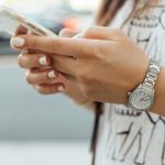 Woman with off-white painted nails holding phone in both hands. Her thumbs are poised over the keys.