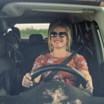 Woman with blonde hair sitting behind the wheel of a car, buckled in, hands on the wheel,, smiling.
