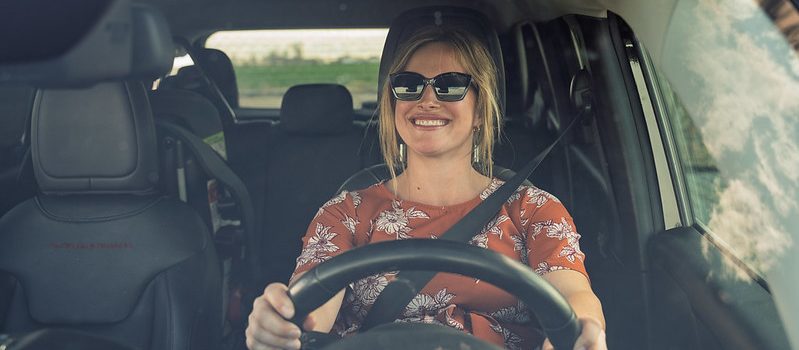 Woman with blonde hair sitting behind the wheel of a car, buckled in, hands on the wheel,, smiling.