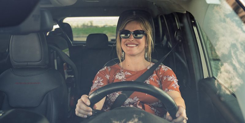 Woman with blonde hair sitting behind the wheel of a car, buckled in, hands on the wheel,, smiling.