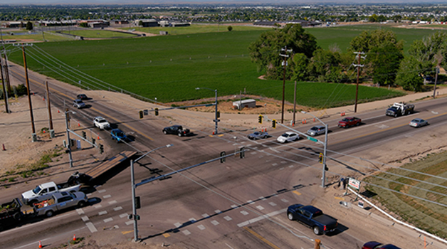 A four lane road intersects with a two lane road in rural area, surrounded by farm fields.