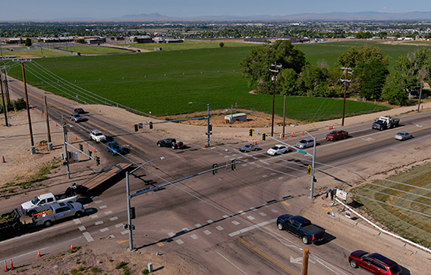 A four lane road intersects with a two lane road in rural area, surrounded by farm fields.