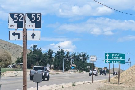 In foreground, state highway signs for both state highway 52 and 55. Behind the signs several vehicles are on a road in a mountain area.