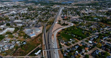 Aerial view of a major highway interchange under construction in an urban area. Vehicles travel along the main freeway while new ramps and overpasses take shape. Surrounding the interchange are residential neighborhoods, commercial buildings, and small industrial facilities. Farther in the distance, rivers, ponds, and open fields stretch toward the horizon under clear skies.