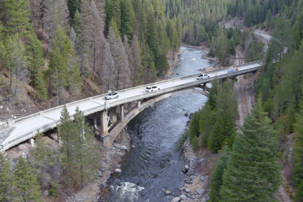 An arch bridge with several cars traversing a river surrounded by pine trees. Bridge shows wear.