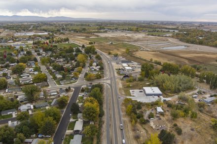 Aerial view of a two-lane highway running through a small town surrounded by houses, trees, and open farmland. Vehicles travel along the road as it curves past neighborhoods and commercial buildings. Beyond the developed area, large fields and ponds stretch toward the horizon under an overcast sky, with low mountains visible in the distance.