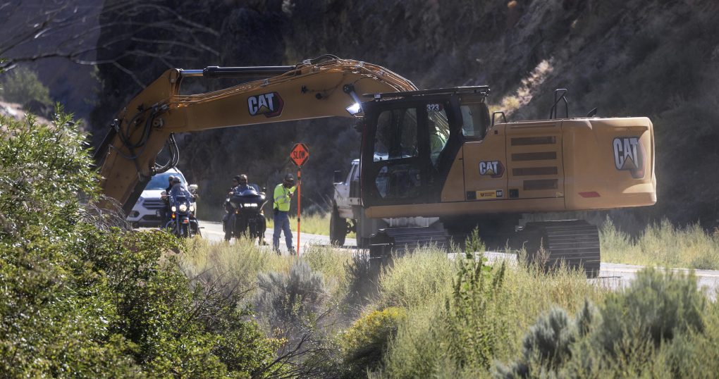 A backhoe on the side of a road. A flagger stops traffic behind it.