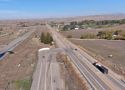 Aerial view of a rural highway with light traffic running through open fields and rolling hills. The main road extends into the distance, intersecting with an older, partially closed off ramp in the foreground. A few homes, trees, and farm buildings are scattered across the landscape under a clear blue sky.