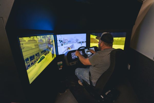 A man wearing a baseball cap and gray shirt sits in a full-scale driving simulator with three large screens displaying a virtual roadway. He grips the steering wheel, focused on the simulation environment showing traffic and buildings.