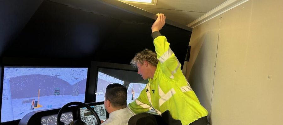 A person wearing a high-visibility yellow jacket leans over the shoulder of another person seated at a driving simulator. The simulator screens display a snowy road scene as the standing person provides instruction or guidance.