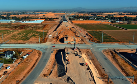Aerial view of an active highway construction site with multiple excavators, loaders, and work trucks operating on a large dirt area. Crews wearing safety vests work near a newly installed concrete box culvert. Construction materials, trailers, and equipment are scattered around the site. In the background, vehicles travel along a nearby highway, and autumn-colored trees and residential buildings border the work zone under clear blue skies.