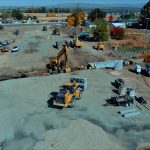 Aerial view of an active highway construction site with multiple excavators, loaders, and work trucks operating on a large dirt area. Crews wearing safety vests work near a newly installed concrete box culvert. Construction materials, trailers, and equipment are scattered around the site. In the background, vehicles travel along a nearby highway, and autumn-colored trees and residential buildings border the work zone under clear blue skies.