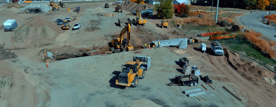 Aerial view of an active highway construction site with multiple excavators, loaders, and work trucks operating on a large dirt area. Crews wearing safety vests work near a newly installed concrete box culvert. Construction materials, trailers, and equipment are scattered around the site. In the background, vehicles travel along a nearby highway, and autumn-colored trees and residential buildings border the work zone under clear blue skies.