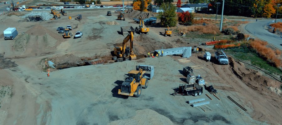 Aerial view of an active highway construction site with multiple excavators, loaders, and work trucks operating on a large dirt area. Crews wearing safety vests work near a newly installed concrete box culvert. Construction materials, trailers, and equipment are scattered around the site. In the background, vehicles travel along a nearby highway, and autumn-colored trees and residential buildings border the work zone under clear blue skies.