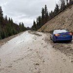 A highway is partially covered in mud and debris from a recent slide. A blue car is stuck in the muddy roadway while a worker in a high-visibility jacket looks on from the side. The scene is surrounded by burned trees and forested hills under an overcast sky.