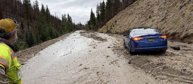 A highway is partially covered in mud and debris from a recent slide. A blue car is stuck in the muddy roadway while a worker in a high-visibility jacket looks on from the side. The scene is surrounded by burned trees and forested hills under an overcast sky.