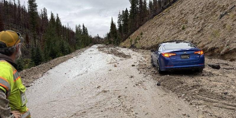 A highway is partially covered in mud and debris from a recent slide. A blue car is stuck in the muddy roadway while a worker in a high-visibility jacket looks on from the side. The scene is surrounded by burned trees and forested hills under an overcast sky.