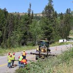 Workers from ITD and IDOC doing hazardous tree removal on SH-31, showing a loader hauling large logs and people on the ground on the roadway around the equipment.