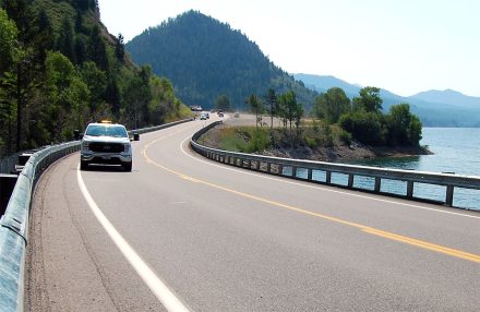 A curving section of US-26 beside Palisades Reservoir in Idaho, showing guardrail and vehicles on the road.