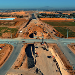 Aerial view of a large highway construction project cutting through open farmland. The image shows a major intersection where crews are building an overpass or interchange, with heavy equipment, cranes, and dirt embankments visible on both sides. Traffic cones line the crossroad, and nearby fields and a few buildings stretch into the distance under a clear blue sky.