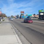 View looking down a busy commercial street with cars driving in both directions. A sidewalk runs alongside the road with fall-colored trees. Visible businesses include Arby’s on the left and Subway on the right. The sky is clear and blue.