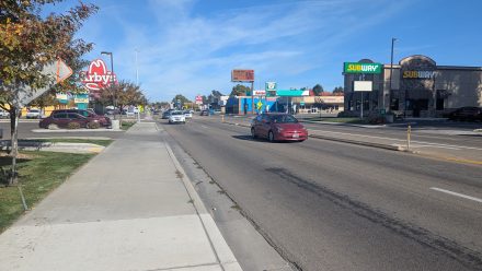 View looking down a busy commercial street with cars driving in both directions. A sidewalk runs alongside the road with fall-colored trees. Visible businesses include Arby’s on the left and Subway on the right. The sky is clear and blue.