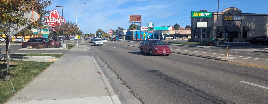 View looking down a busy commercial street with cars driving in both directions. A sidewalk runs alongside the road with fall-colored trees. Visible businesses include Arby’s on the left and Subway on the right. The sky is clear and blue.