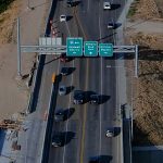 Aerial view of traffic traveling across a bridge on State Highway 55 in Nampa, passing under overhead signs for I-84 west to Caldwell and Ontario, Midland Boulevard north, and the Karcher Bypass.