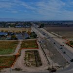 Aerial view of a roadway under construction running through farmland and developed areas, with vehicles traveling on the open lanes and work zones marked by orange fencing on both sides of the corridor.