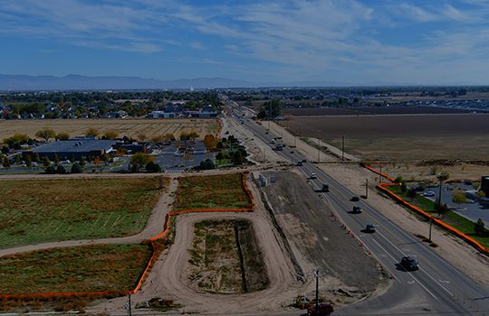Aerial view of a roadway under construction running through farmland and developed areas, with vehicles traveling on the open lanes and work zones marked by orange fencing on both sides of the corridor.