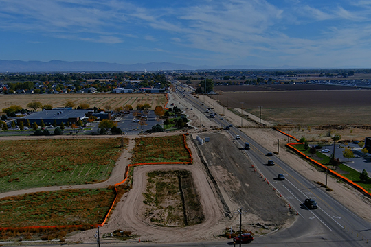 Aerial view of a roadway under construction running through farmland and developed areas, with vehicles traveling on the open lanes and work zones marked by orange fencing on both sides of the corridor.