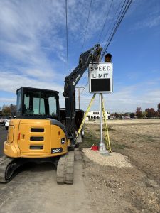Construction equipment installs a variable speed limit sign along a roadway, with the speed display panel visible.
