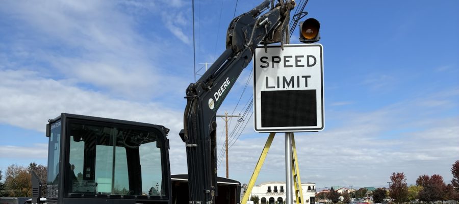 Construction equipment installs a variable speed limit sign along a roadway, with the speed display panel visible.