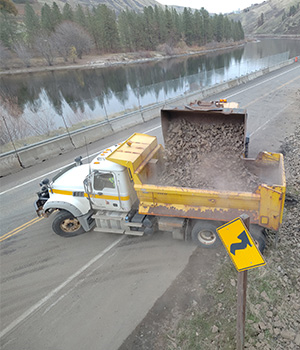 On the US-12 emergency slope project near Arrow, a loader empties a bucket of rocky debris over a truck bed.